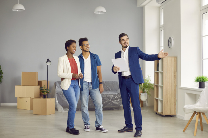 A professional consultant gesturing toward a bright, open living space for a young couple, illustrating what are real estate advisory services in the context of home buying and property selection.